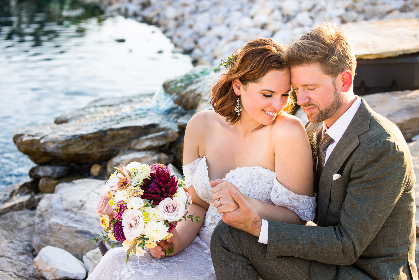image of the couple as they sit down against the rocks and the pond while holding hands and putting their heads together as the bride holds flowers by iron willow floral designs