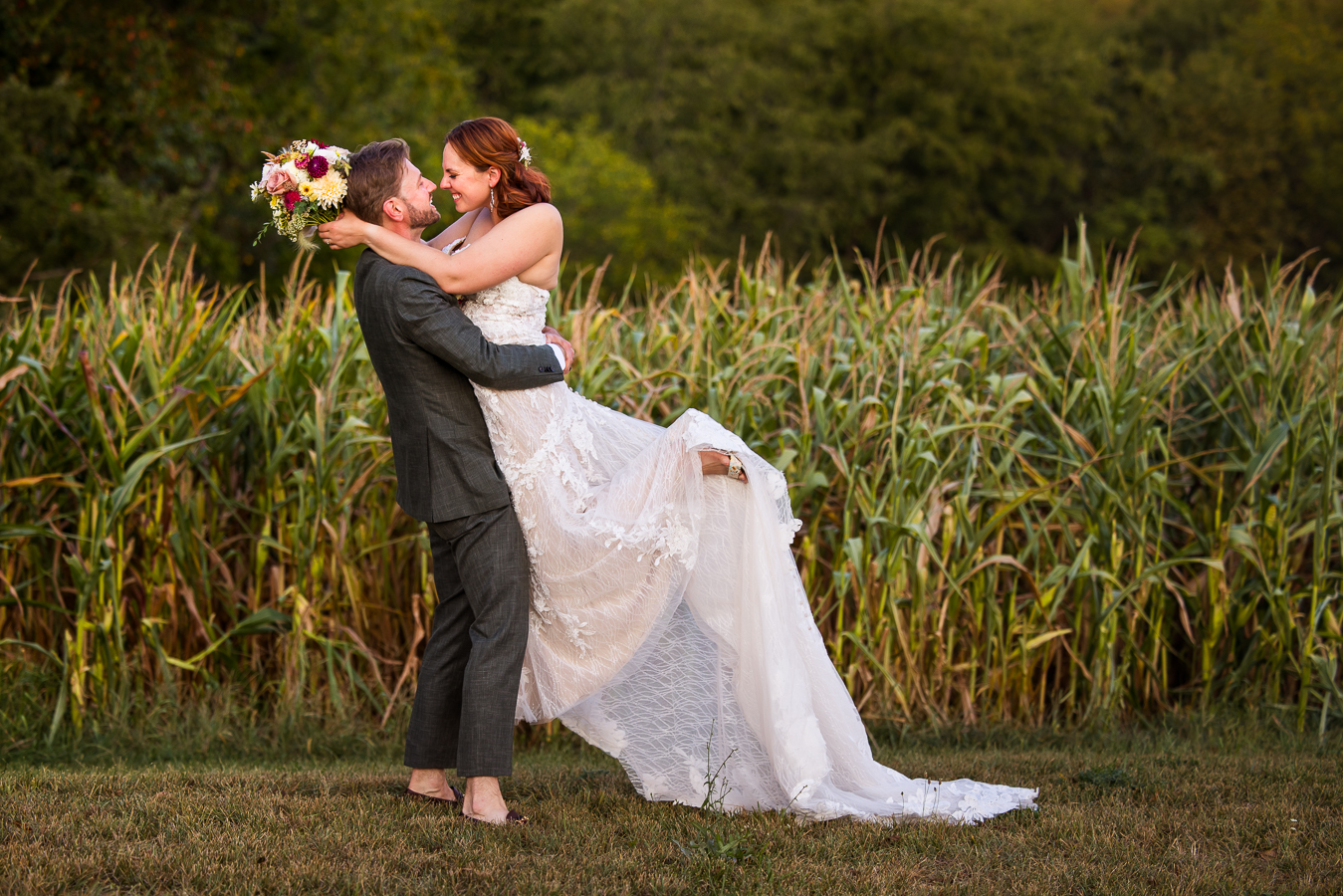 pa wedding photographer, lisa rhinehart, captures this image of the bride and groom he lifts the bride up and they look into each others eyes as they stand against the corn field behind them at this warfordsburg pa wedding venue, alpine acres