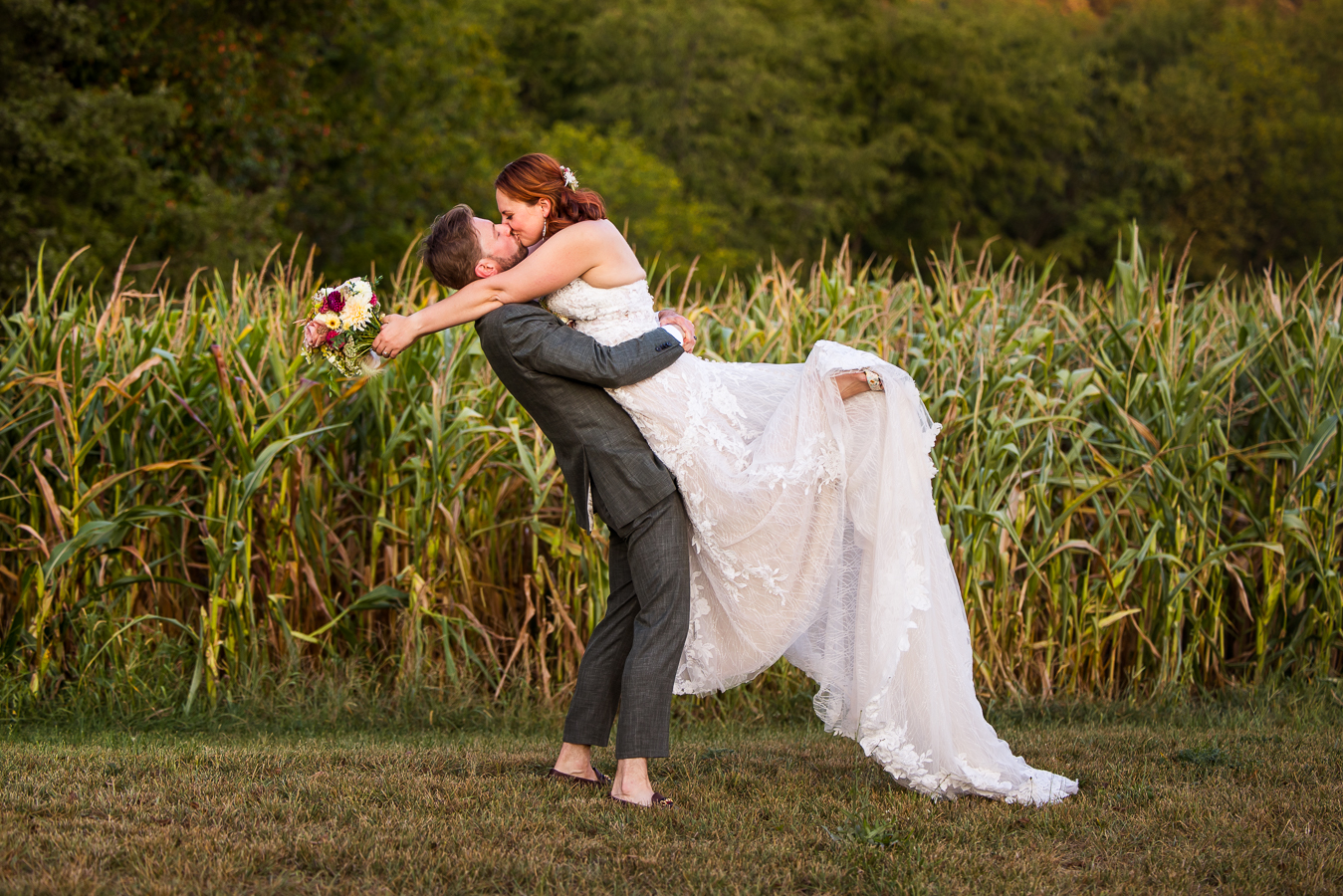 pa wedding photographer, lisa rhinehart, captures this image of the bride and groom he lifts the bride up and kiss one another as they stand against the corn field behind them at this warfordsburg pa wedding venue, alpine acres
