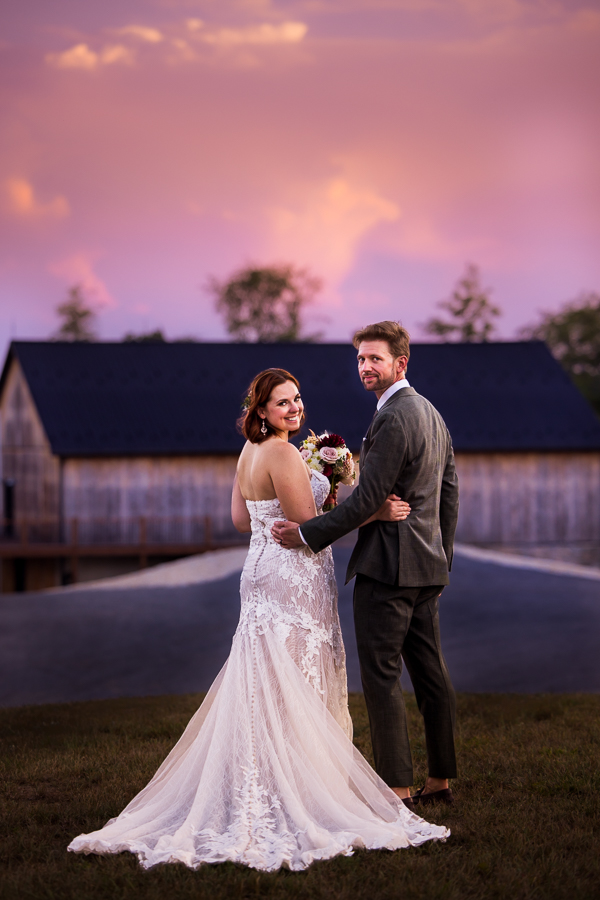 image of this couple as they stand in front of the barn with their hands behind each others back and smiling towards the camera with a vibrant stunning pink, purple sunset behind them 