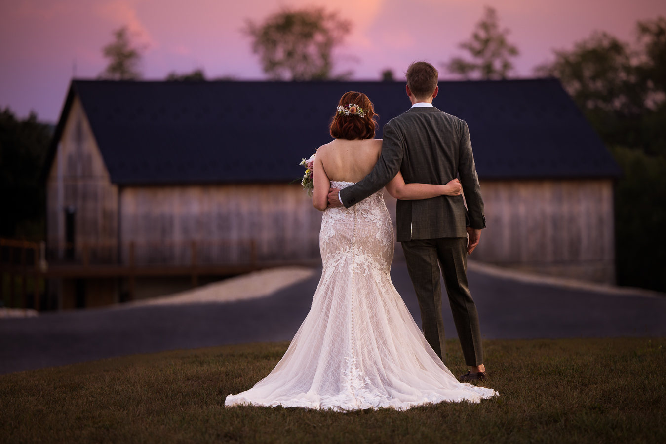 image of this couple as they stand with their backs to the camera with their arms around each other facing the rustic barn with a vibrant pink, purple sunset as they explore alpine acres one of Central PA Wedding Venues