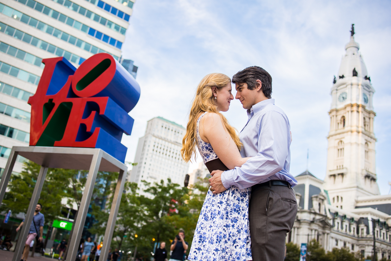 creative, unique image of the couple as they standing face one another hugging and smiling with the giant love sign and other Philadelphia landmarks behind them for this creative philly engagement 