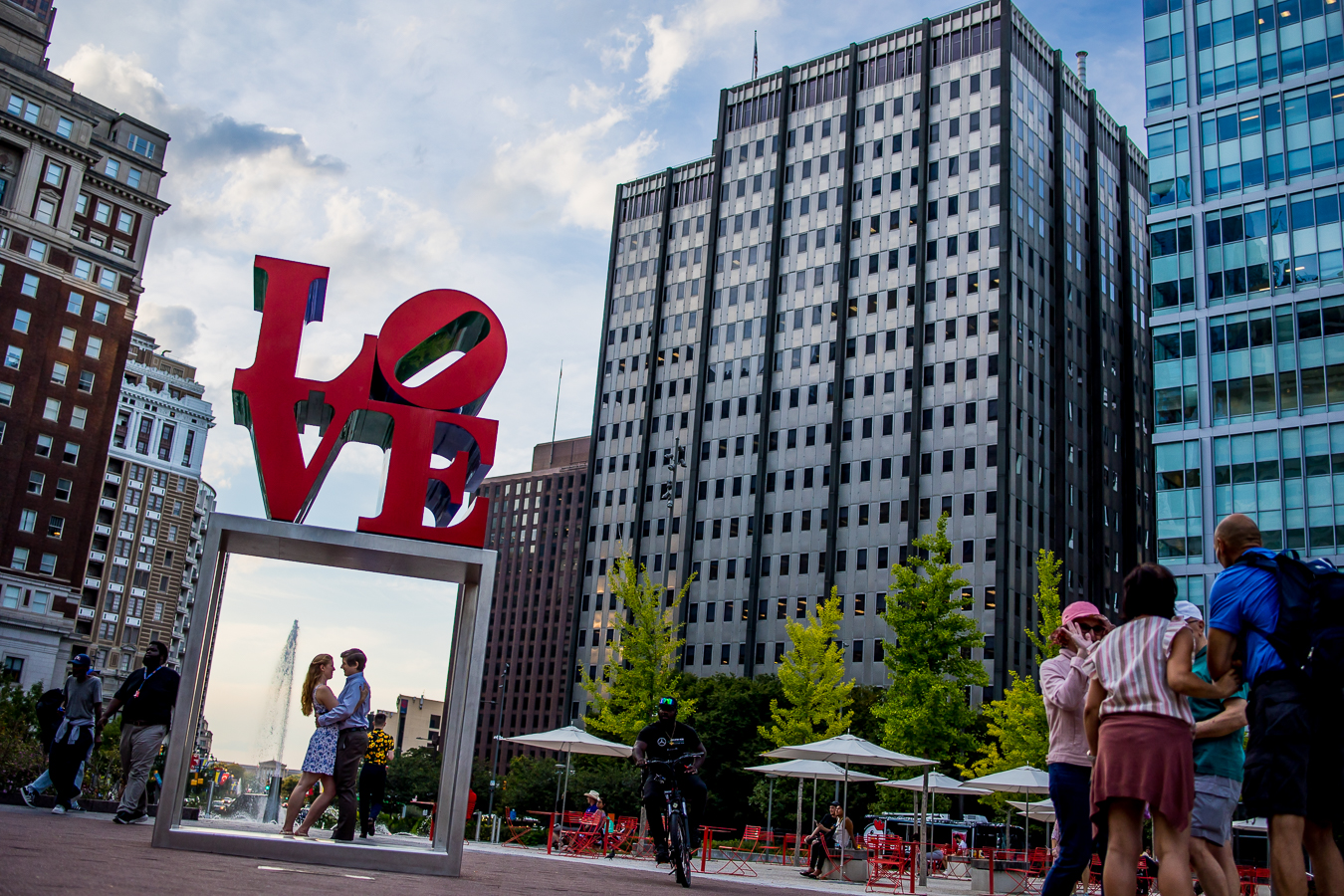 Creative Photojournalistic Philadelphia Wedding Photographer, rhinehart photography, captures this image of the couple as they stand together underneath the famous love sign in center city Philadelphia while the city buzzes with action around them 