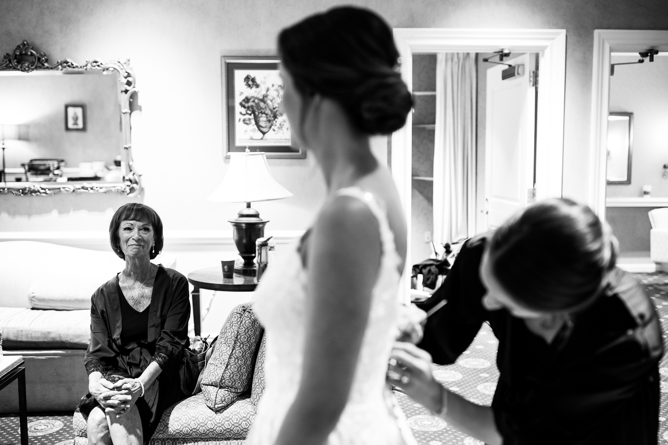 black and white image of the brides mom watching her get dressed in her wedding gown as she smiles at her daughter during the wedding preparations at the palace at somerset park captured by NJ wedding photographer, lisa rhinehart