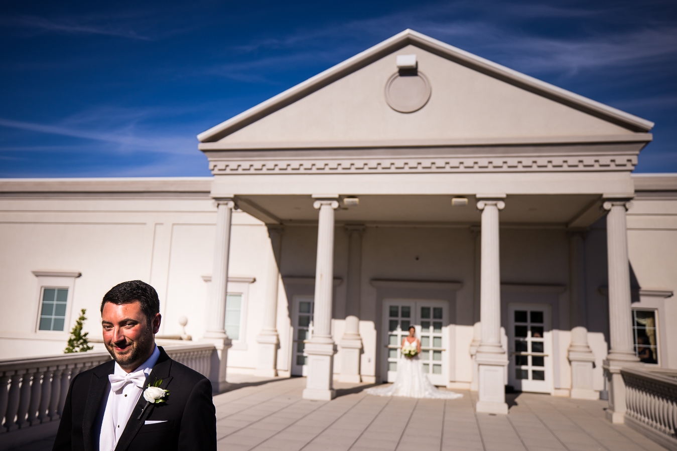 luxury nj wedding photographer, lisa rhinehart, captures this vibrant image of the groom smiling as his bride stands behind him in her wedding gown as they get ready to see each other for the first time during their first look outside of the palace at somerset park 