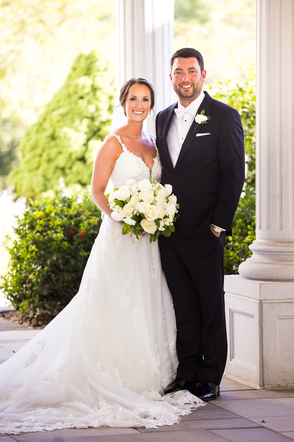 the palace wedding photographer, lisa rhinehart, captures this traditional portrait of the bride and groom as they stand beside one another smiling at the camera outside of the palace at somerset park in front of the large white columns and vibrant green trees