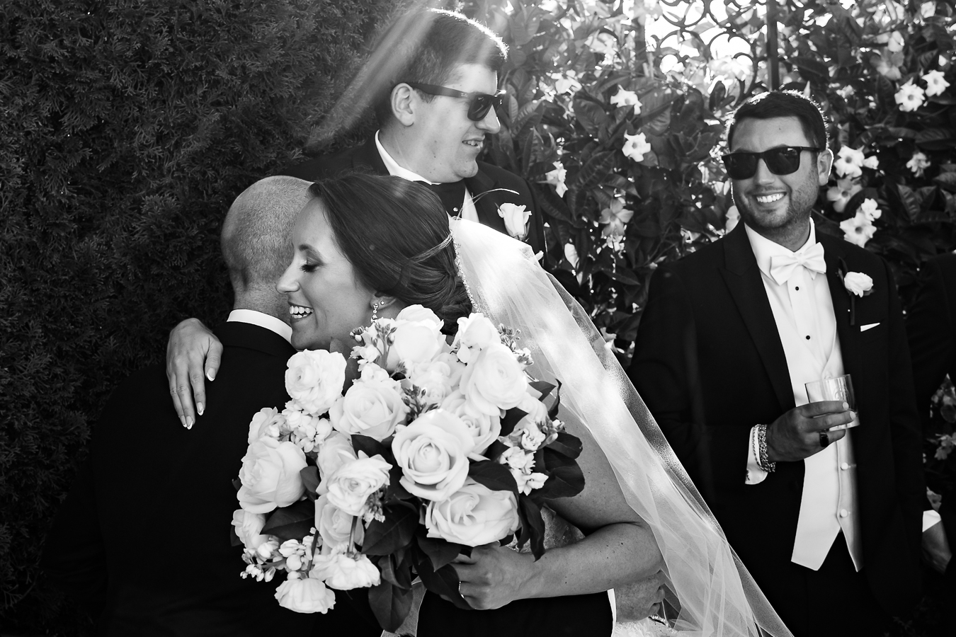 candid nj wedding photographer, lisa rhinehart, captures this black and white candid image of the bride as she hugs some groomsmen during the wedding party photos outside of the palace in NJ 