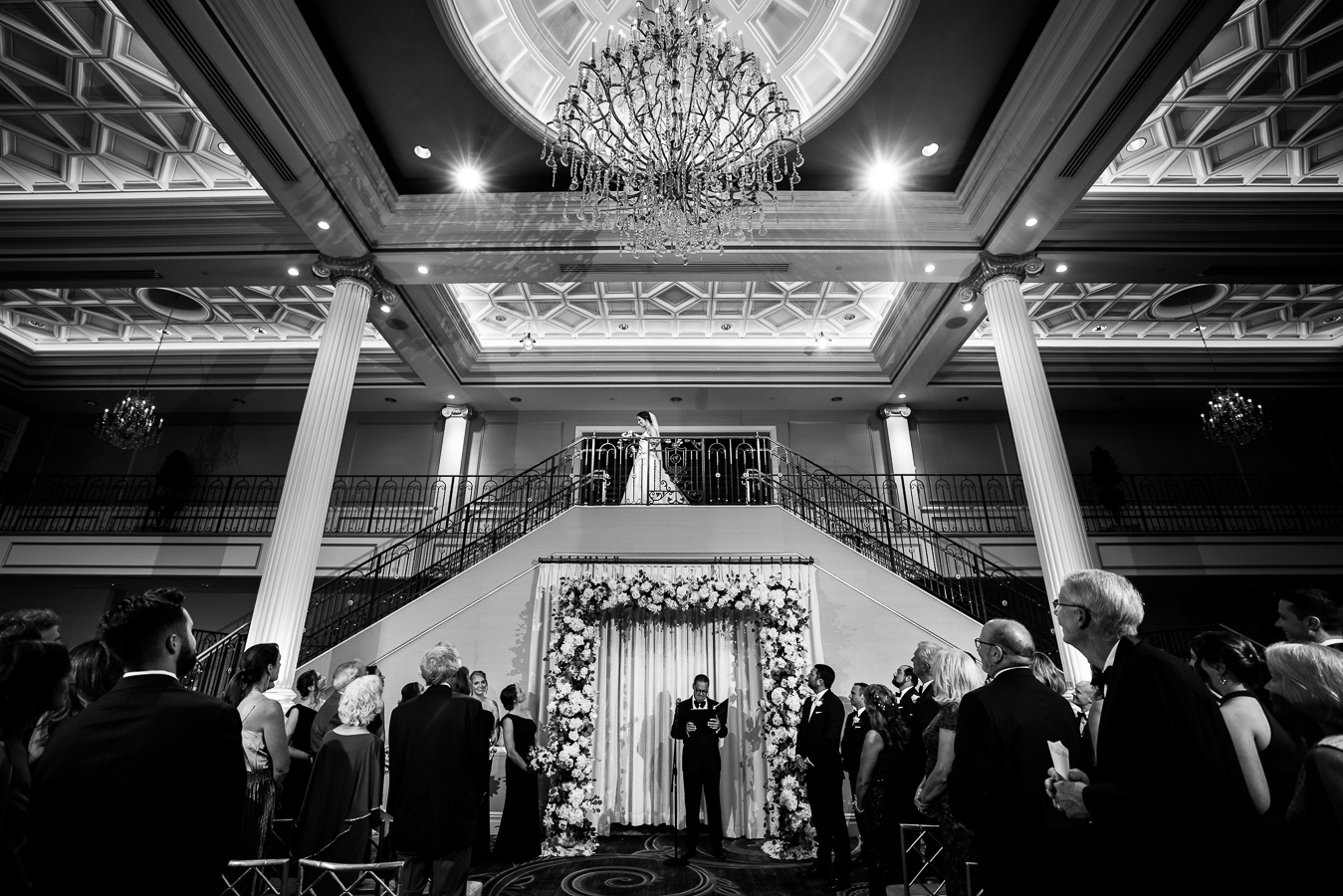 unique, creative perspective of the ceremony space and guests as they watch the bride walk down the grand staircase in the black and white moment captured by creative nj palace wedding photographer, lisa rhinehart