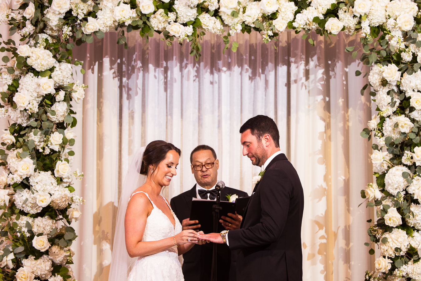 the palace wedding photographer, lisa rhinehart, captures this image of the bride and groom as they exchange their vows and rings with a giant white floral arch in the background behind them during this indoor palace wedding ceremony in NJ 