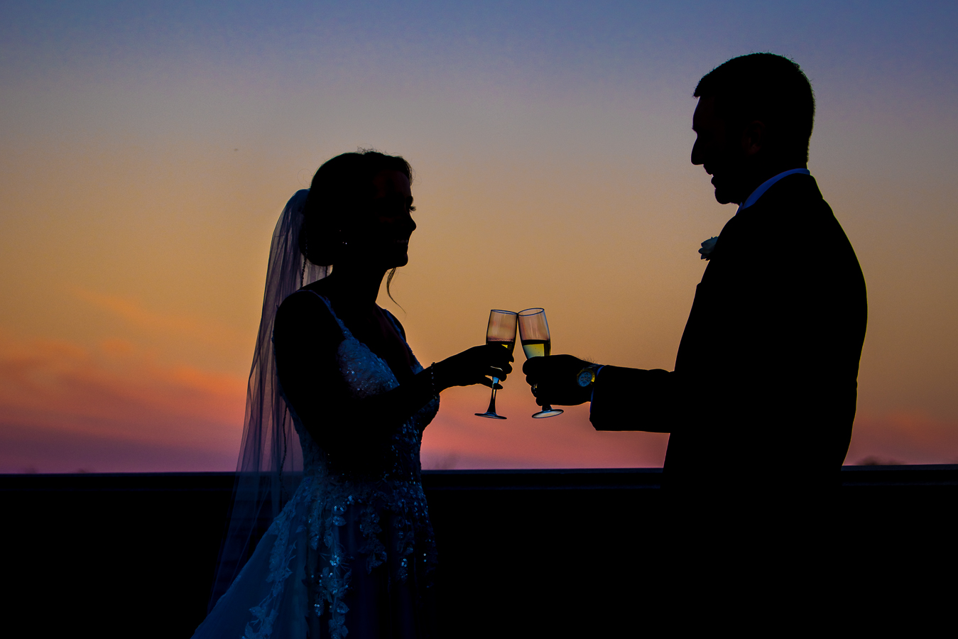 creative nj wedding photographer, lisa rhinehart, captures this unique, creative silhouetted image of the bride and groom as they share a drink while the vibrant, colorful sunset is captured behind them during their romantic portrait session before their black and gold wedding reception at the palace at somerset park in new jersey 