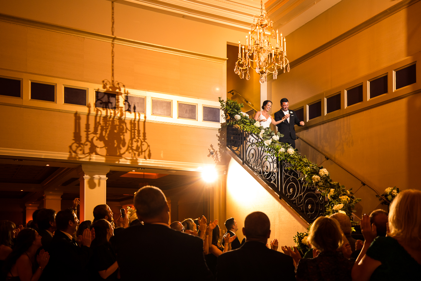nj wedding photographer, lisa rhinehart, captures this black and gold wedding reception moment of the bride and groom as they walk down the grand staircase holding hands into their wedding reception at the palace at somerset park 