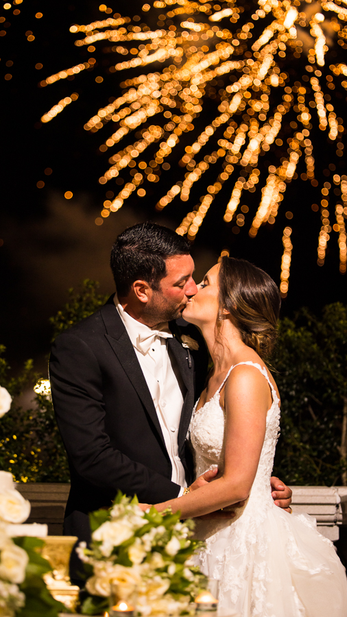 nj wedding photographer, lisa rhinehart, captures this black and gold wedding theme at the palace at somerset park as this newlywed couple shares a kiss together as gold fireworks are set off in the background behind them 