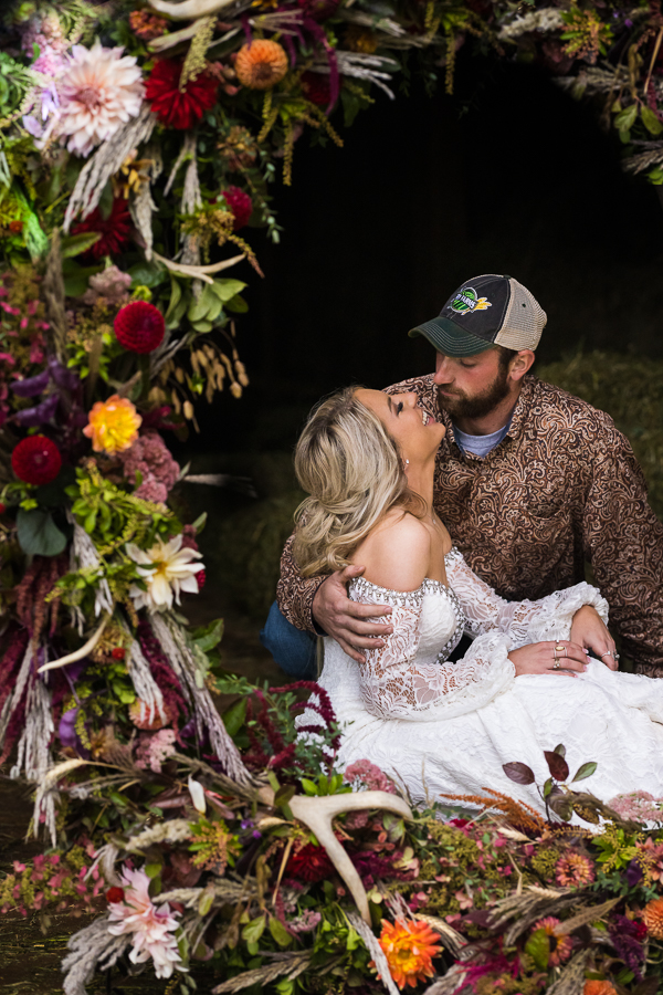 The bride and groom sitting on the ground of the barn looking into each others eyes as the vibrant colorful flower decor and antlers fill the frame around them during this outdoor, barn wild country wedding