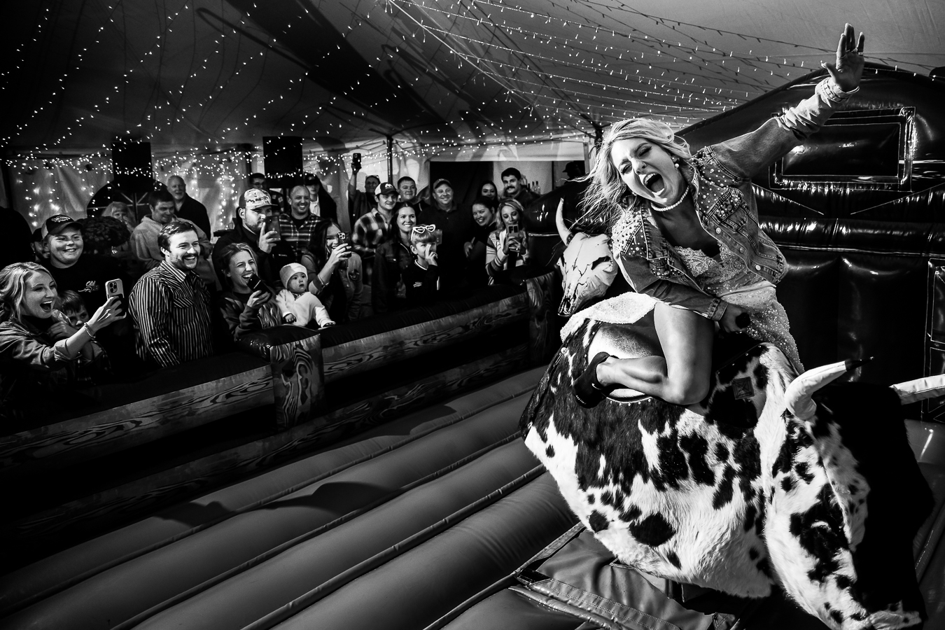 fun, candid black-and-white image of the bride as she rides the mechanical bull at her wild country wedding as her guests cheer for her during this fun, unique country wedding reception
