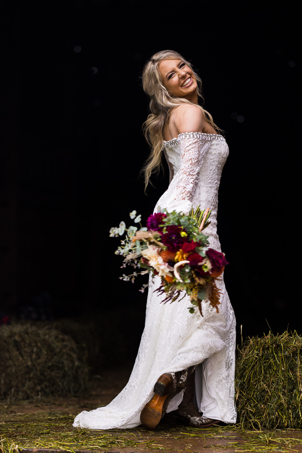 country wedding photographer, lisa rhinehart, captures this fun, unique bridal portrait of this country bride as she stands in the barn holding her floral bouquet showing off her boots and smiling back at the camera 
