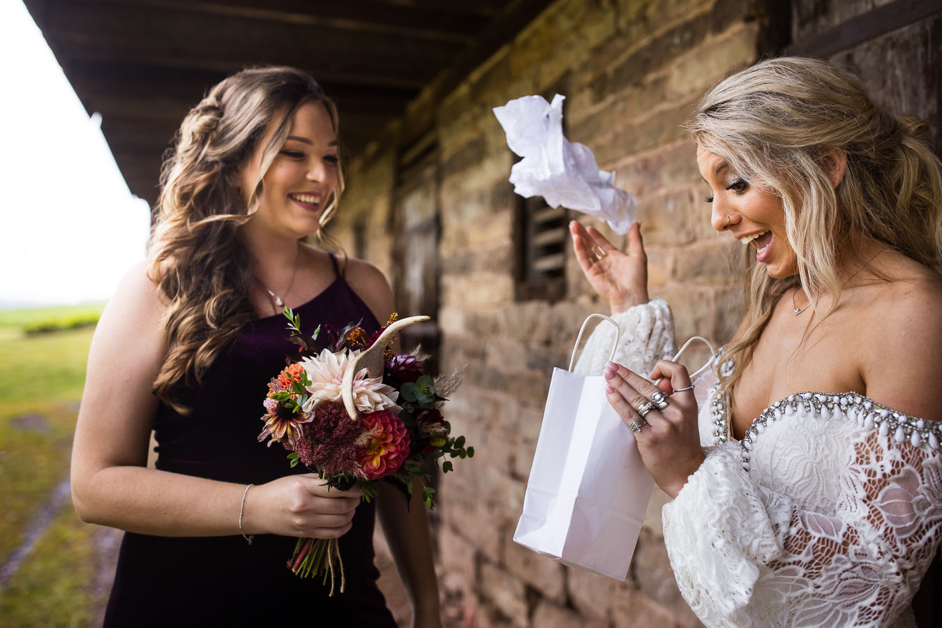 fun, candid image of the bride as she opens a gift from her bridesmaid and looks happy and surprised as she throws the tissue paper before her outdoor wedding ceremony 