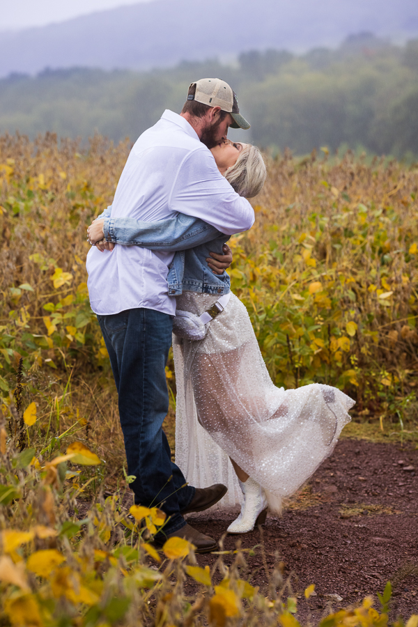 pa wedding photographer, lisa rhinehart, captures the newlyweds as they kiss each other out in the middle of the field surrounded by vibrant yellow colored crops and the mountains behind them during their country inspo wedding reception 