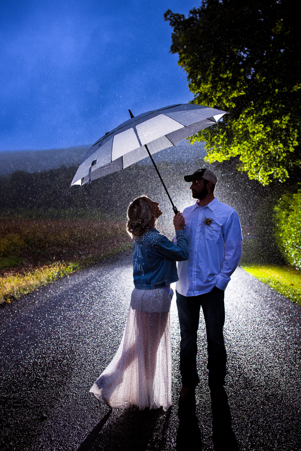 creative pa country wedding photographer, rhinehart photography, captures the bride and groom as they stand with each other on the road underneath the umbrella during a downpour at their country inspired wedding in halifax pa 