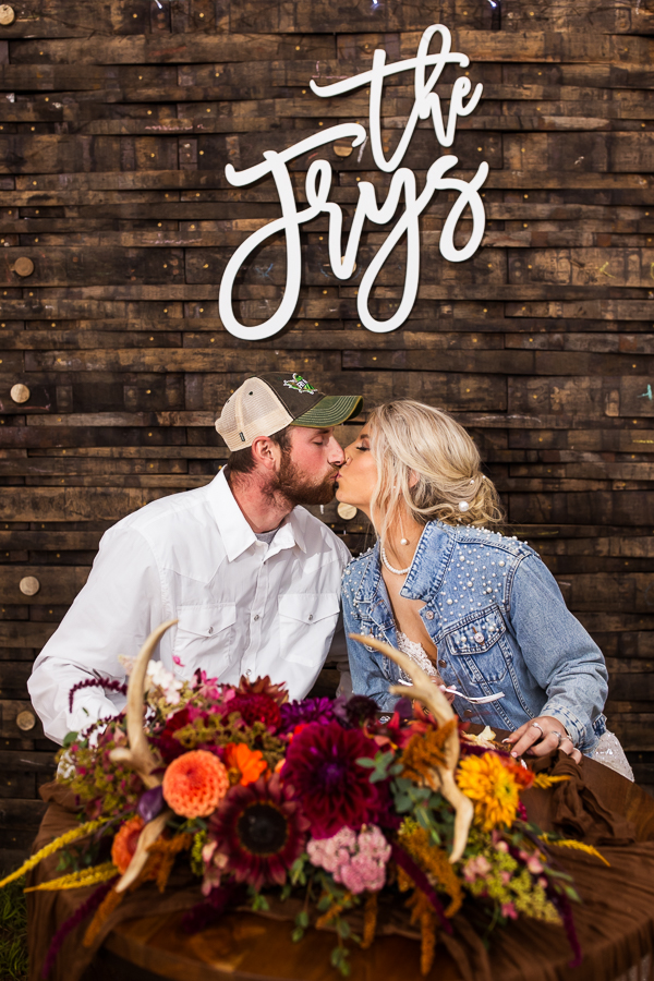 wild country wedding photographer, lisa rhinehart, captures the bride and groom as they share a kiss together underneath their unique custom last name sign with their unique vibrant fall florals and antlers on the table in front of them during this country inspo wedding reception 