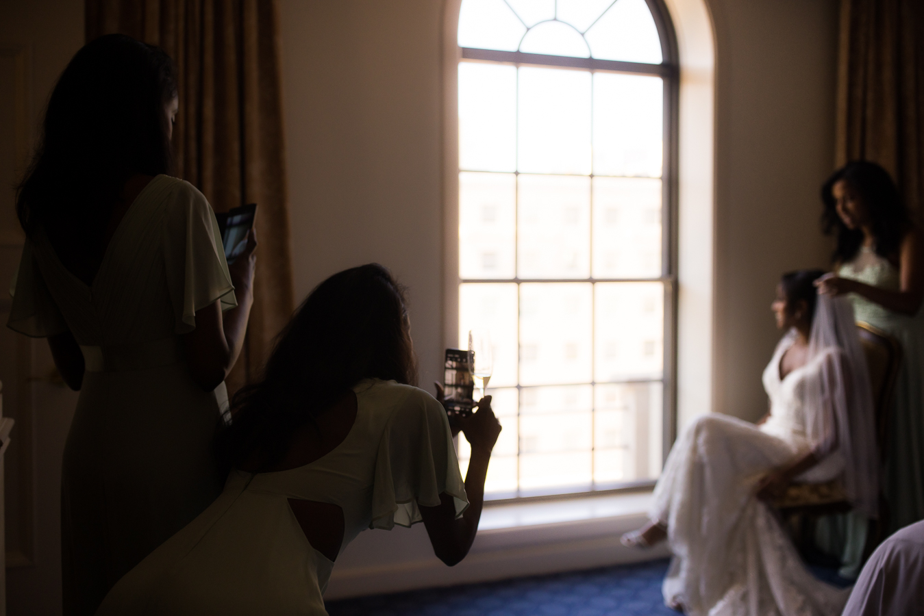 candid image of the bridesmaids capturing shots of the bride as she sits in a chair and gets her veil put on at the st regis hotel in Washington DC