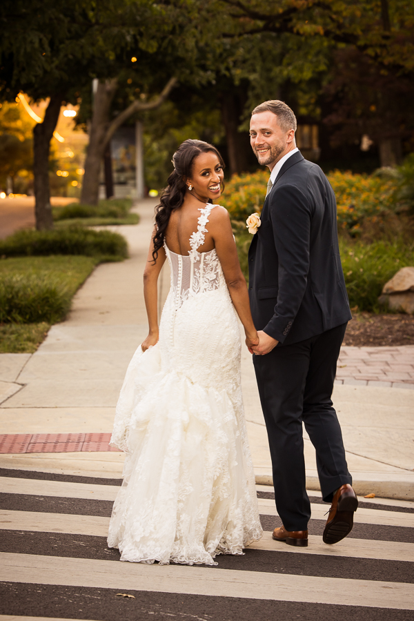 fun image of the bride and groom as they walk hand and hand across the crosswalk in Washington DC as they look back and smile and laugh at the camera during their romantic portrait session during golden hour after their wedding reception at st francis hall 