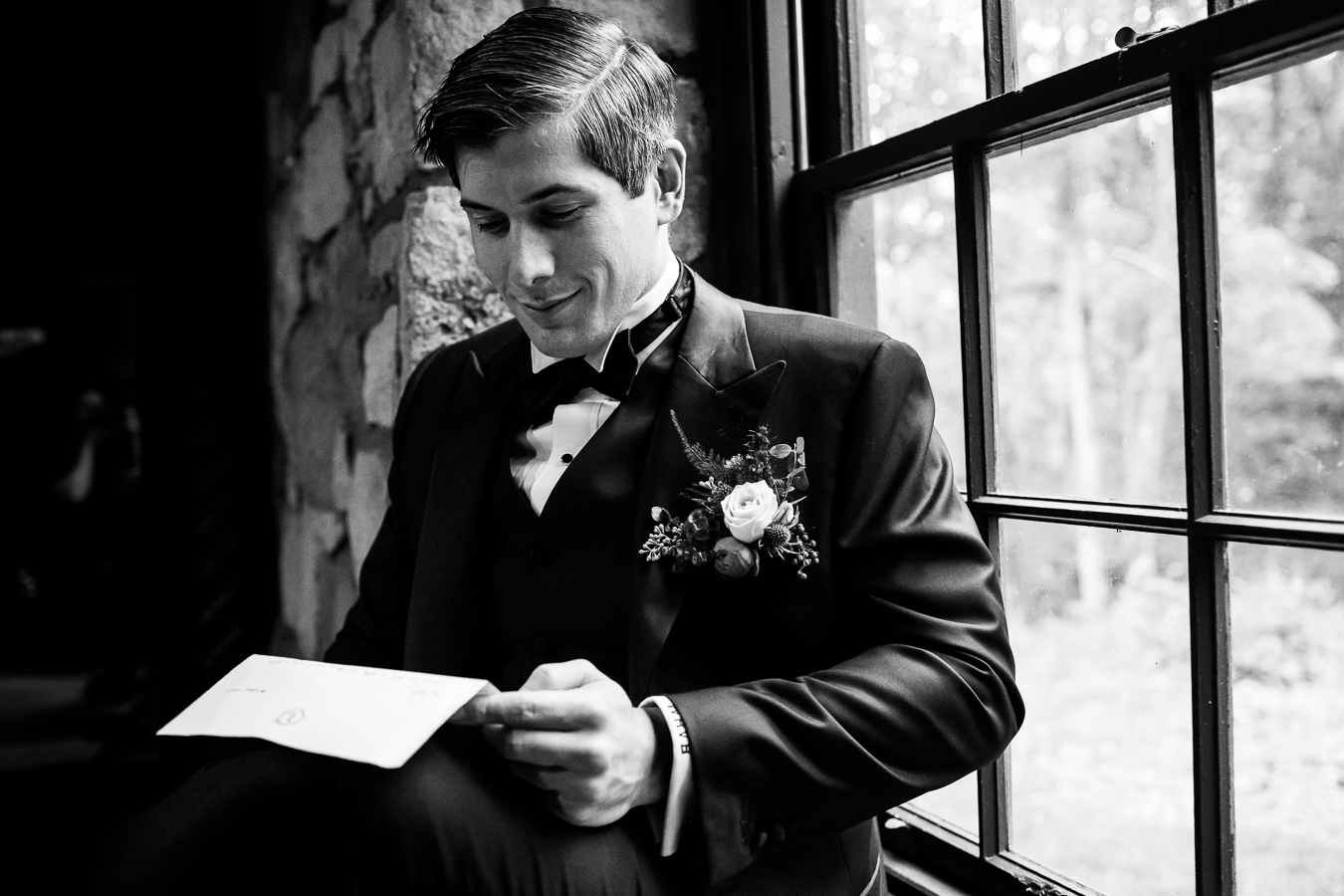 black and white image of the groom as he stands beside a window reading a letter and smiling before seeing his bride for the first time at their Elizabeth Furnace Wedding