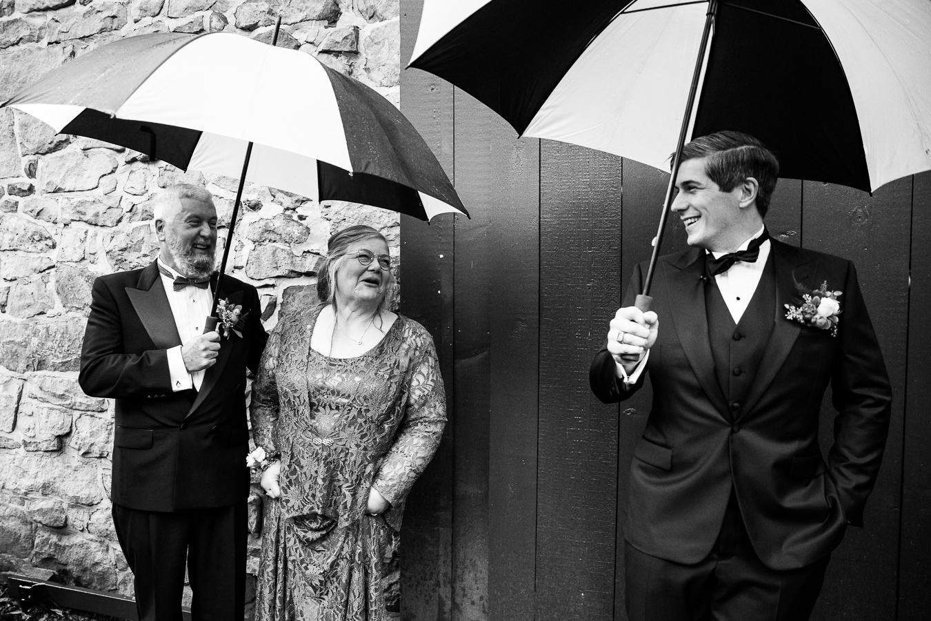 Elizabeth Furnace Wedding photograher, Lisa Rhinehart, captures this black and white candid image of the groom and parents as they stand outside of the ceremony barn with umbrellas over their heads to stay out of the rain 
