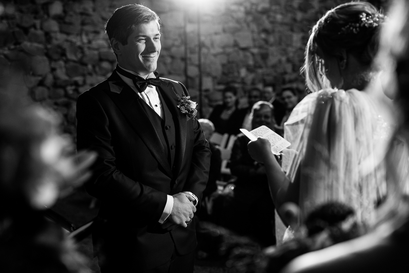 candid black and white moment between the bride and groom as they share their vows as the groom smiles big at his bride during their Elizabeth Furnace Wedding ceremony 