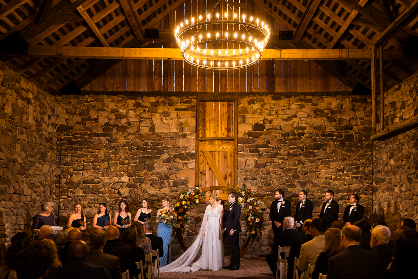 image of the bride and groom as they stand at the end of the aisle with their wedding party on both sides of them and the giant chandelier over top of them during this indoor wedding ceremony in lititz pa in the charcoal barn