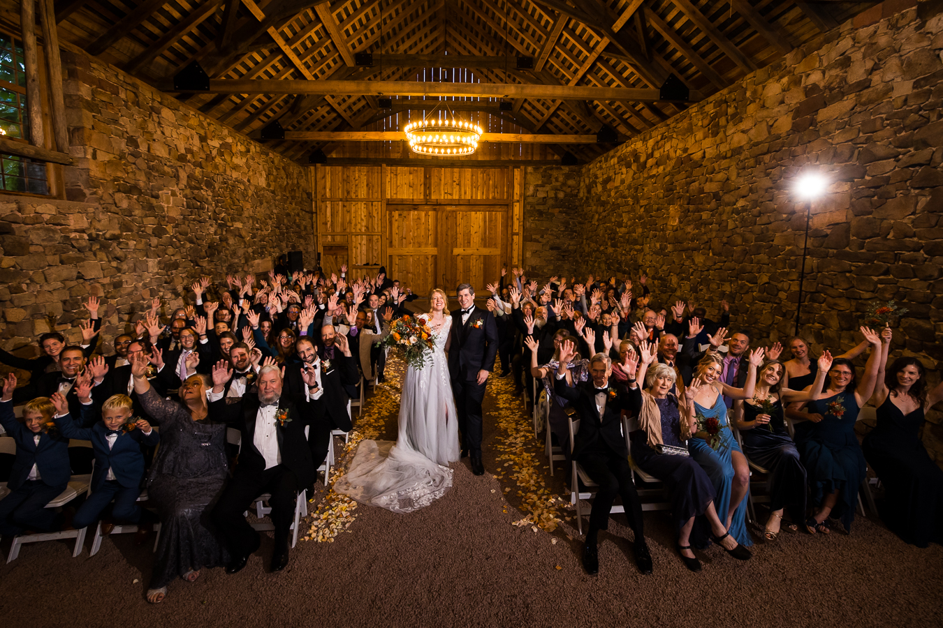 a large group shot of the bride and groom in the middle and all of their guests sitting around them inside of the barn at the furnace with their hands up cheering and smiling 