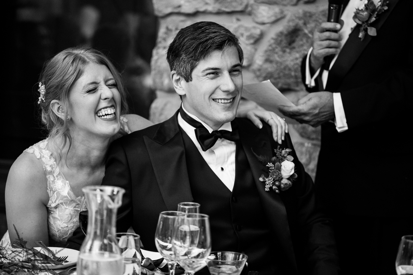 black and white image of the bride and groom as they smiles and listen to their friends and family giving speeches at this fun wedding reception 