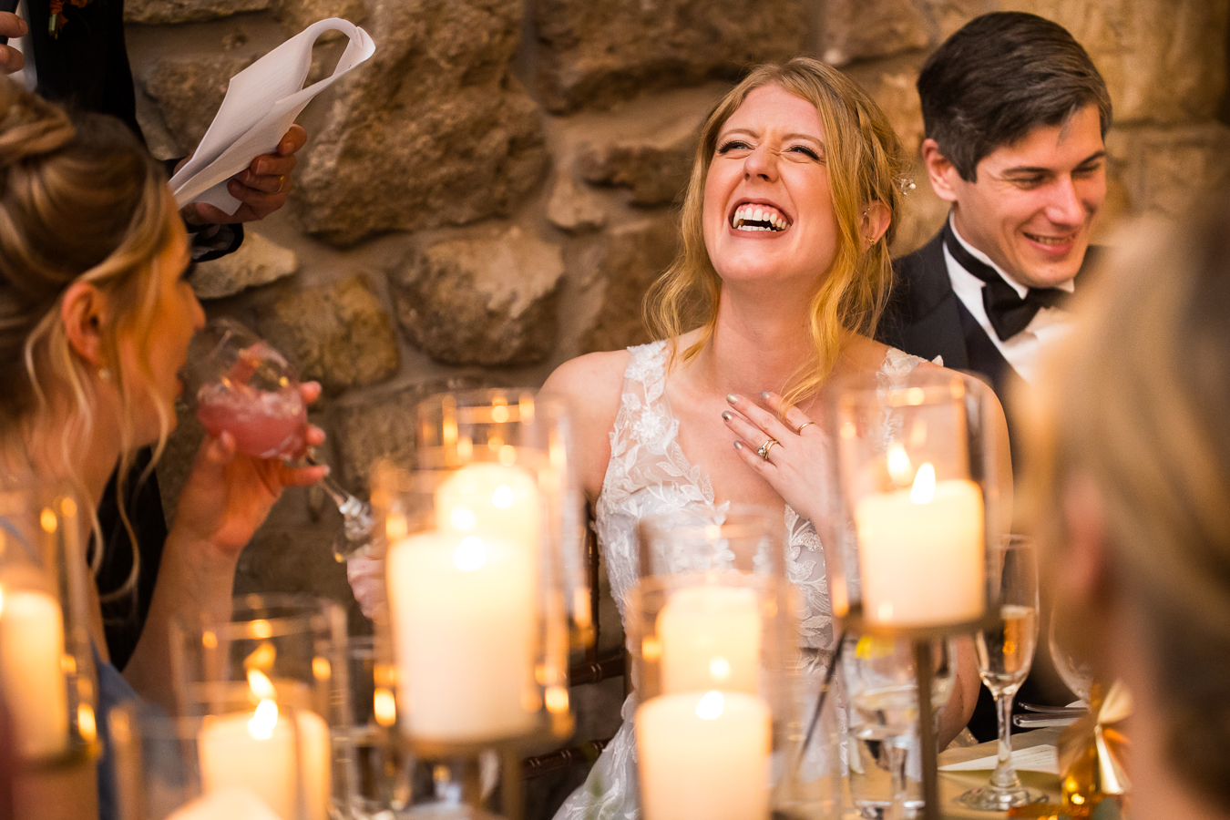 image of the bride as she smiles and laughs with her hand on her chest as her friends and family share their heartfelt speeches during the wedding traditions portion of the reception 
