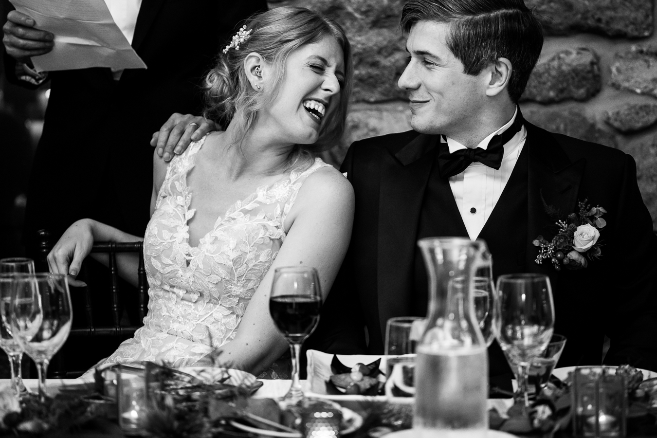 black and white image of the bride and the groom as they lean in towards one another and smile as their families share their speeches at this wedding reception 