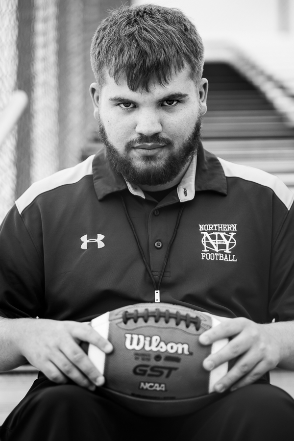 black and white image of this high school football coach as he stares down the camera as if its his opponent while holding a football during this high school portrait session at northern york football stadium 