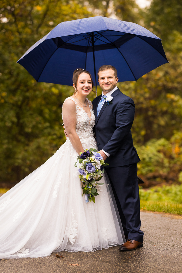 traditional portrait of the bride and groom as they stand next to one another and smile at the camera underneath the umbrella to avoid their rainy day wedding 