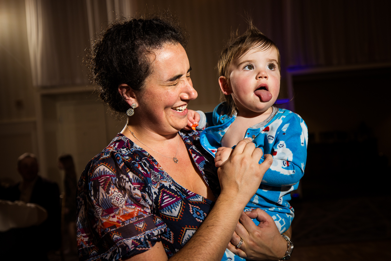 image of a wedding guest who is holding a baby that is sticking their tongue out as the lady laughs during this indoor wedding reception 