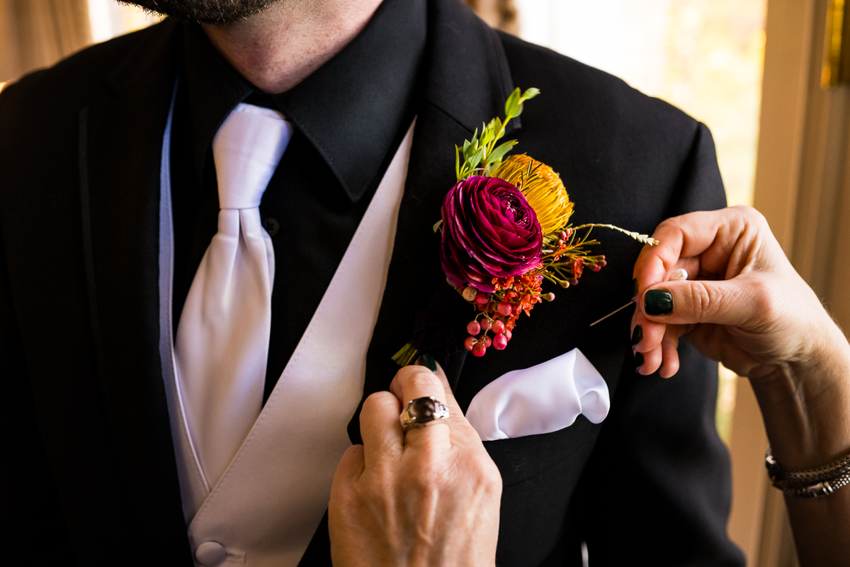 close up image of the groom getting his vibrant colorful boutonniere placed on his black jacket before this outdoor fall wedding ceremony in Leesburg va