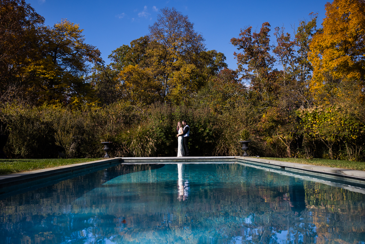 landscape portrait of the bride and groom as they hug one another at the end of the reflecting pool located at Murray hill virgina surrounded by the fall foliage of leesburg virginia 