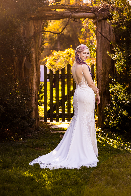 traditional bridal portrait of the bride as she stands in the wooden archway with her back to the camera as she shows off the back of her wedding dress and rainbow hair as she stands outside in Leesburg va 