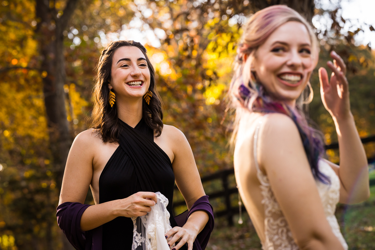 candid image of a bridesmaid as she smiles at the bride with her vibrant rainbow hair as they stand outside surrounded by the vibrant fall foliage of leesburg va