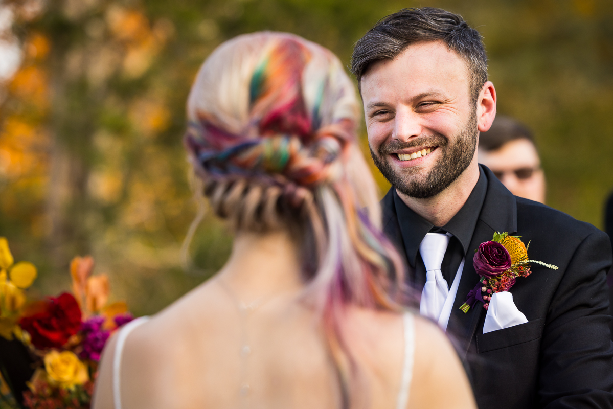 candid portrait of the groom as he is all smiles while looking at his bride and her rainbow hair during their outdoor fall wedding ceremony at Murray hill in Virginia beside the Potomac river 