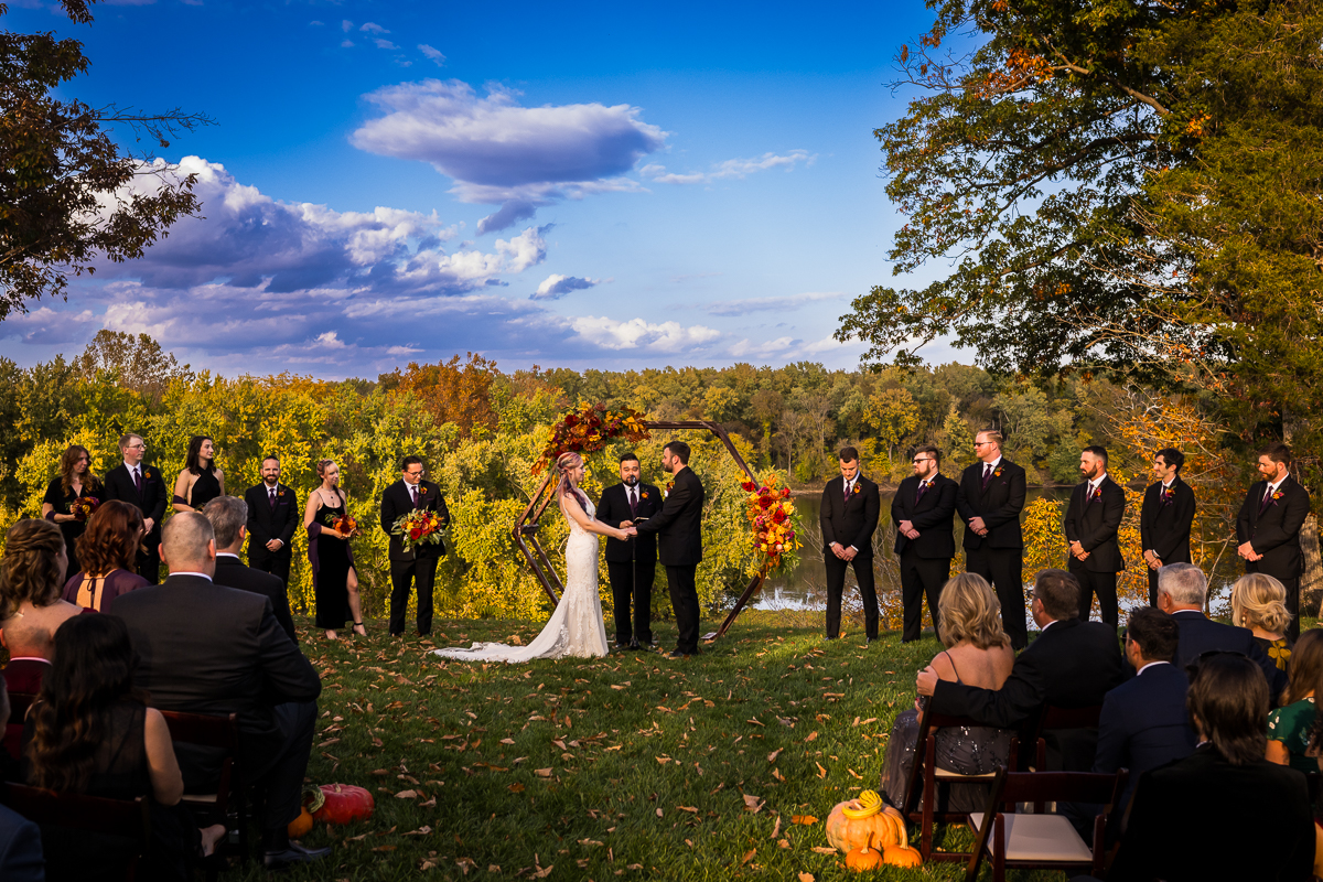 Murray Hill wedding photographer, lisa rhinehart, captures this stunning landscape image of the bride and groom as they hold hands during their outdoor vibrant fall wedding ceremony surrounded by family, friends and vibrant fall foliage 
