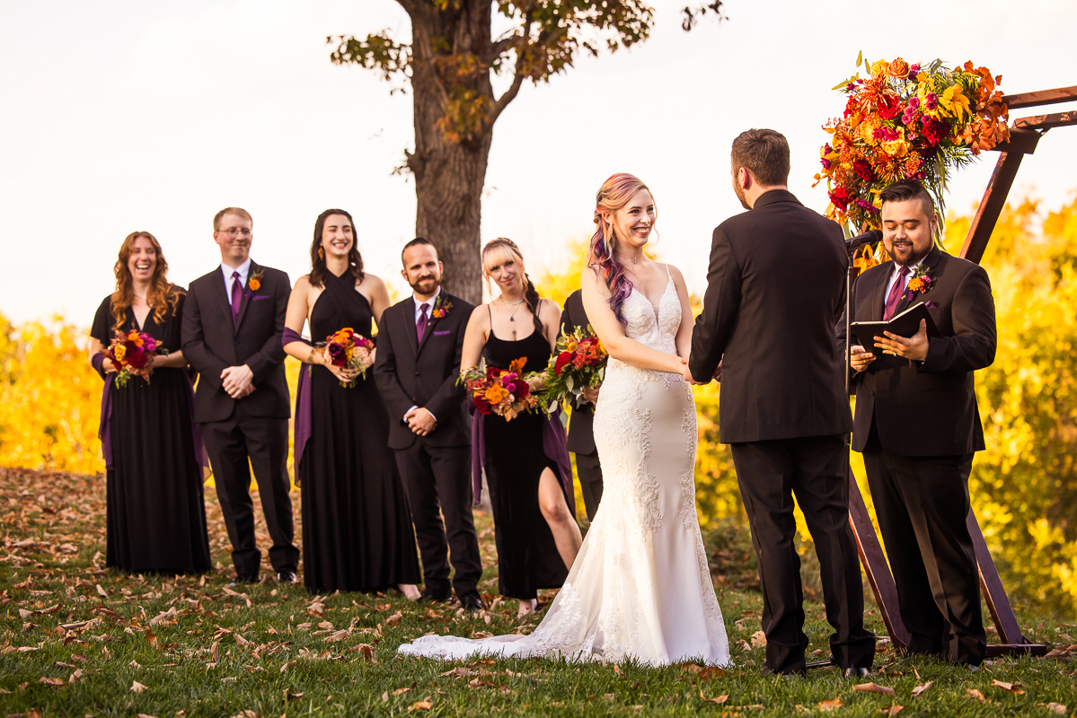 candid wedding photographer, lisa rhinehart, captures this image of the bride and groom as they hold hands, while their wedding party smiles and watches during this vibrant outdoor fall wedding ceremony 