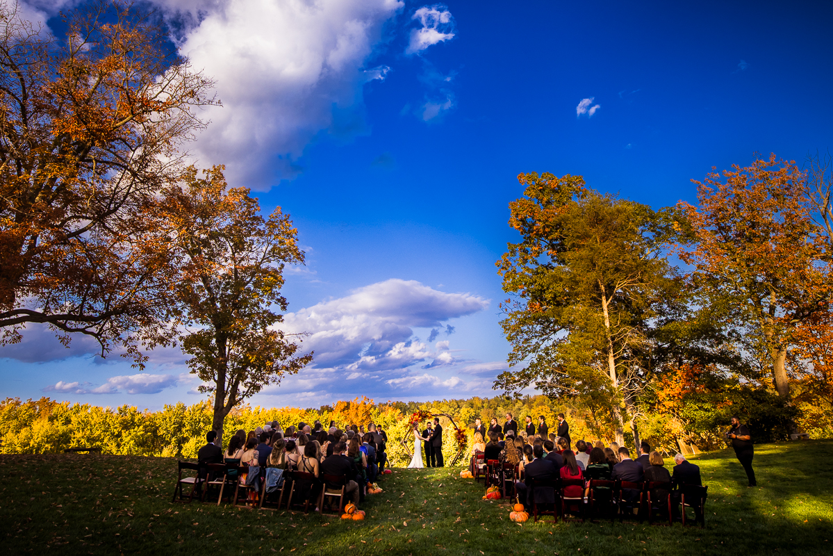 Murray Hill wedding photographer, lisa rhinehart, captures this vibrant, colorful landscape image of the bride and groom holding hands at the end of the aisle surrounded by vibrant fall foliage, friends and family as they watch this wedding ceremony beside the Potomac river