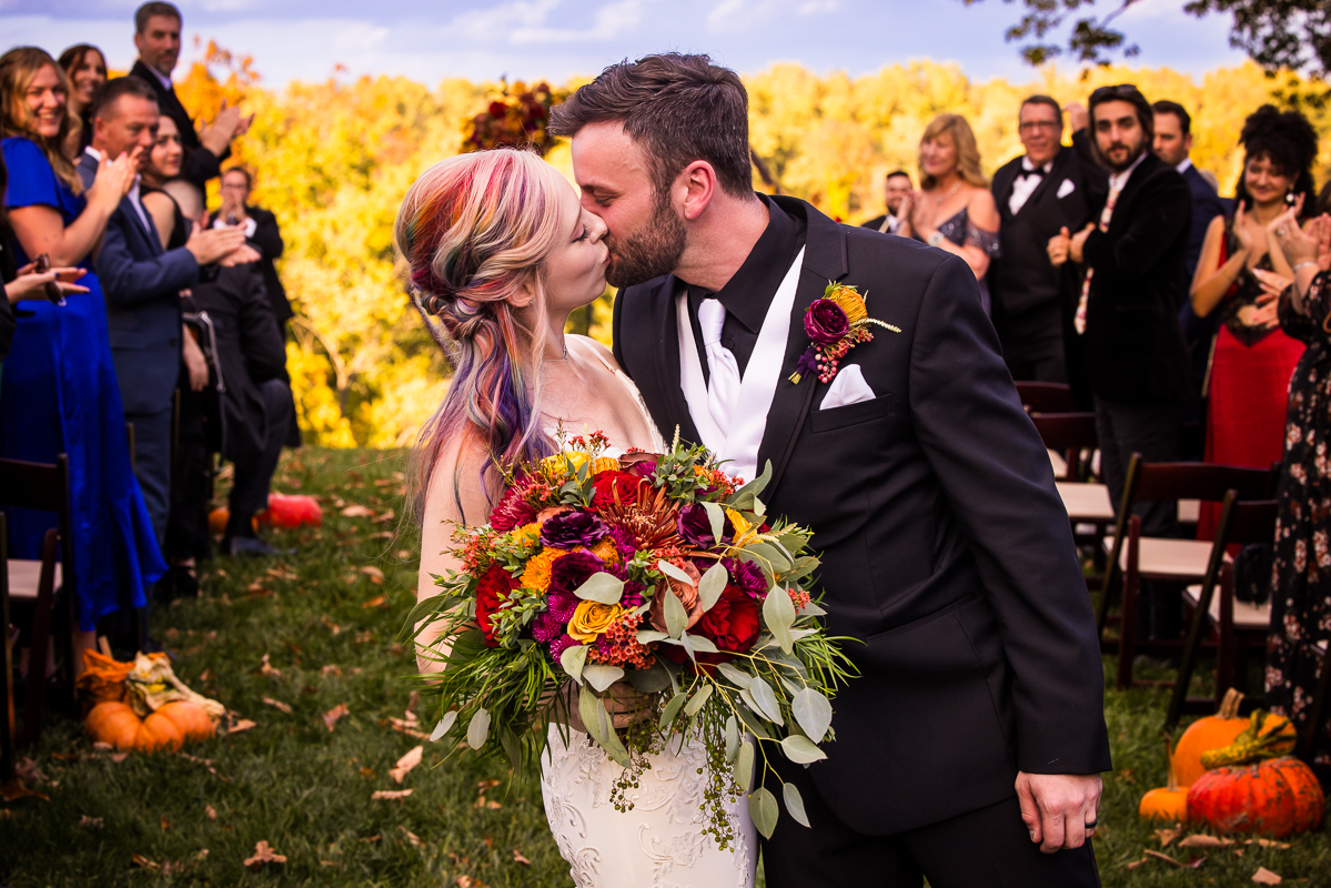 Murray Hill wedding photographer, lisa rhinehart, captures this vibrant, colorful image of the bride and groom as they share a kiss at the end of the aisle while their family and friends cheer and clap behind them surrounded by vibrant colors from the fall foliage and the brides rainbow hair 