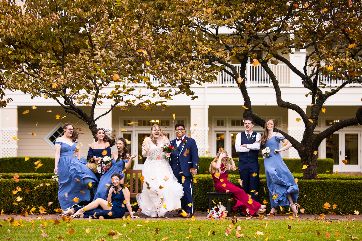 Outdoor vogue shot of the bride and groom with their wedding party laughing and smiling as the colorful fall leaves blow in the wind in front of them in front of the Omni in Bedford captured by creative Omni wedding photographer, lisa rhinehart