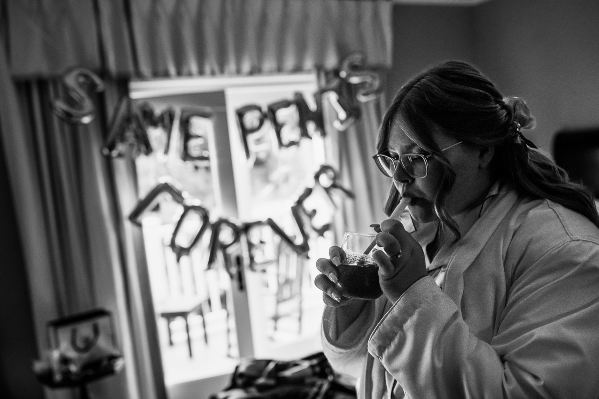 bedford pa wedding photographer, lisa rhinehart, captures this black and white image of the bride as she drinks her drink before her wedding ceremony while getting ready in the suite at omni bedford springs