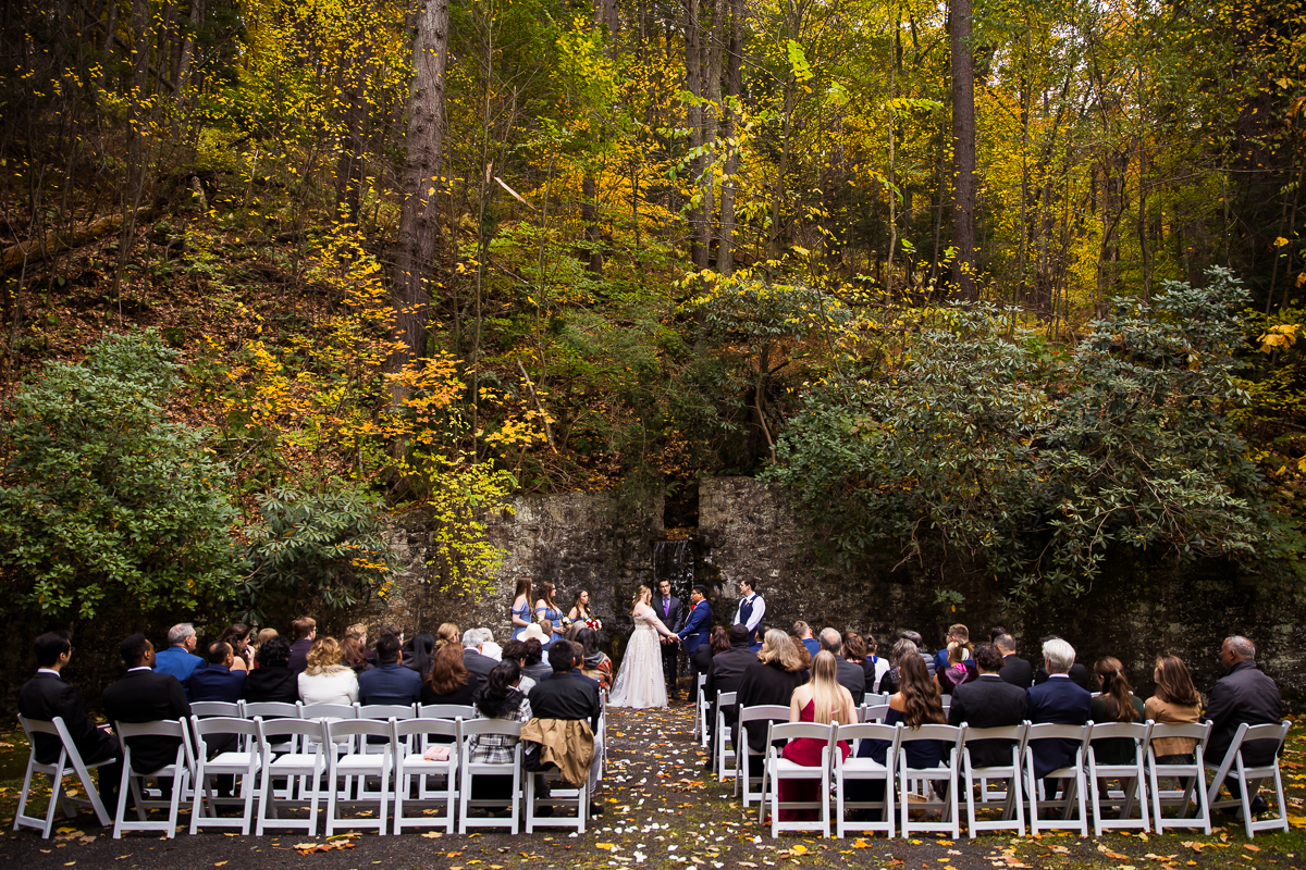 omni bedford springs wedding photographer, lisa rhinehart, captures this landscape image of this outdoor fall wedding ceremony of the bride and groom holding hands at the alter surrounded by friends, family, and vibrant fall foliage in Bedford pa 