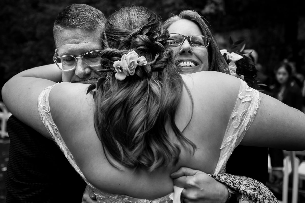 candid, emotional, authentic black and white image of the bride as she hugs her mom and dad after this outdoor fall wedding ceremony at omni bedford springs in Bedford pa 