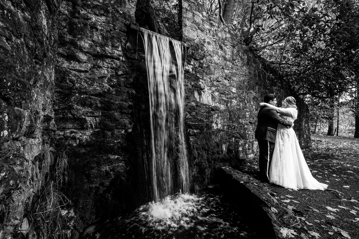 creative Omni wedding photographer, lisa rhinehart, captures this black and white image of the bride and groom as they hug one another and smile while standing by the waterfall located at omni bedford springs in Bedford pa 